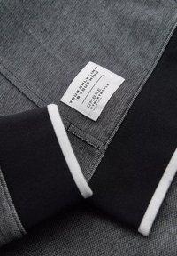 Tejido gris texturizado con un collar negro que presenta acentos de franjas blancas. Incluye una etiqueta con texto y nombre de la marca.