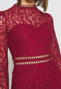 Burgundy spetsklänning med långa ärmar, intrikata blommönster, hög halsringning och cirkulär utskärningsdetalj vid midjan.
