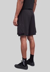 Shorts de sport noirs avec une taille élastique, texture lisse et une poche cargo sur le côté. Portés avec des chaussures et des chaussettes noires.