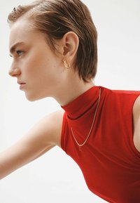 Top sin mangas rojo con cuello alto, acompañado de pendientes de aro dorados y un delicado collar de oro, centrado en la textura y el detalle.