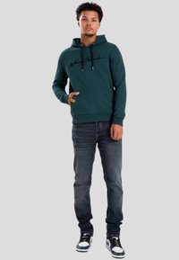 Felpa verde con cappuccio e testo nero, tasca frontale e etichetta arancione. Abbinata a jeans scuri e sneakers bianco e nere.
