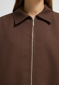 Brun stofjakke med krave, med en sølvfarvet lynlås fortil. Materialet har en tekstureret, ribbet overflade.