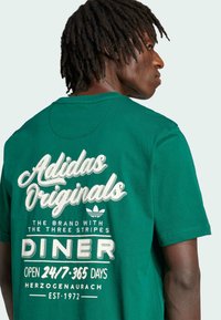 Green cotton t-shirt featuring large white printed text that reads "Adidas Originals," "Diner," and "Open 24/7/365 Days," with a crew neckline.