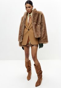 Bruine nepbont oversized jas over een beige op maat gemaakte blazer, groene shirt en bruine shorts. Bruine slouchy kniehoge laarzen maken de look af.