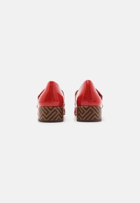 Pantofi din piele patent roșie cu toc jos, decorati cu un model în zigzag de culoare maro pe toc. Textură netedă, design cu vârful ascuțit.