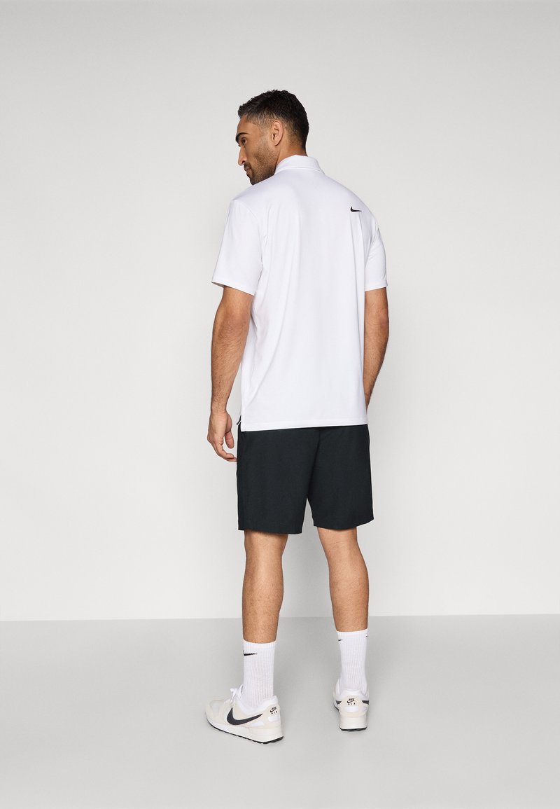 Polo blanc avec un petit col et des détails noirs, associé à un short noir. Le mannequin porte des baskets blanches avec des accents noirs.