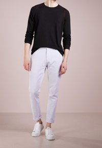 Persona con una camicia nera a maniche lunghe, pantaloni grigi chiari arrotolati e sneakers bianche, in piedi contro uno sfondo chiaro e uniforme.