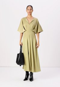 Rochie maxi verde măsliniu cu mâneci scurte și largi și decolteu adânc în V. O pereche de cizme negre cu toc înalt și o geantă tote neagră. Textură netedă.