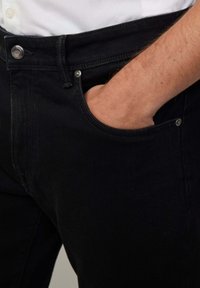 Crne traperice od denima imaju prednju džep, metalne detalje i glatku teksturu, a u njima je dio ruke koji se odmara u džepu.