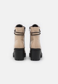 Bottines en cuir verni beige avec semelles en caoutchouc noires, dotées d'une fermeture éclair, de détails texturés et de deux sangles élastiques noires.