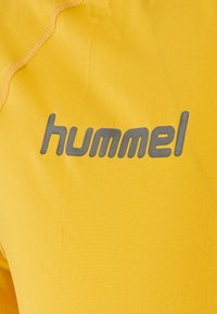 Gul atletisk tyg med en slät textur som har en framträdande grå "hummel"-logotyp. Designen är minimalistisk med en ren finish och subtila stygn.