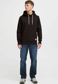 Sudadera negra con un bolsillo tipo canguro y cordones blancos, combinada con jeans azules y zapatillas grises. Diseño simple, sin patrones ni acentos.