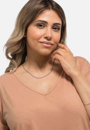 Silberne Kettenhalskette, die am Hals getragen wird, mit einem dünnen, filigranen Design und einer glatten, glänzenden Oberfläche gegen ein beiges V-Ausschnitt-Shirt.
