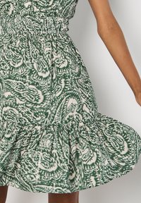 Grün-weiß gemustertes Kleid mit tailliertem Schnitt, ausgestellt Rock und Rüsche am Saum. Hergestellt aus leichtem Stoff mit strukturierten Details.