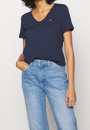 Henkilö, jolla on yllään tummansininen V-kaula-aukollinen lyhythihainen paita, joka on tungettu vaaleansinisiin korkeavyötäröisiin farkkuihin, ja toisessa kädessä taskussa.