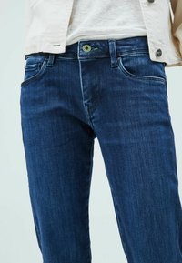 Detailní záběr osoby v tmavě modrých džínách a béžové bundě přes bílou košili, ukazující přední kapsy a detaily knoflíků.