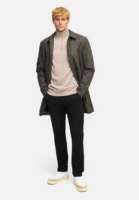 Olivgrön lättviktsjacka över en beige zip-up-tröja, kombinerad med svarta byxor och krämfärgade sneakers med bruna accenter.