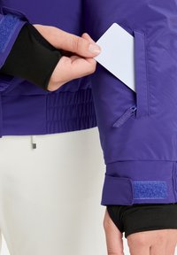 Purppurainen eristetty takki, jossa on vetoketjullinen tasku hihassa ja kontrastinen musta sisäkangasta. Takin pinnassa on sileä rakenne ja tarranauhatehosteita.