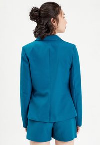 Ensemble blazer et shorts en teal, design ajusté, texture lisse, col à crans, fente arrière unique et shorts à revers. Aucun détail matériel visible.