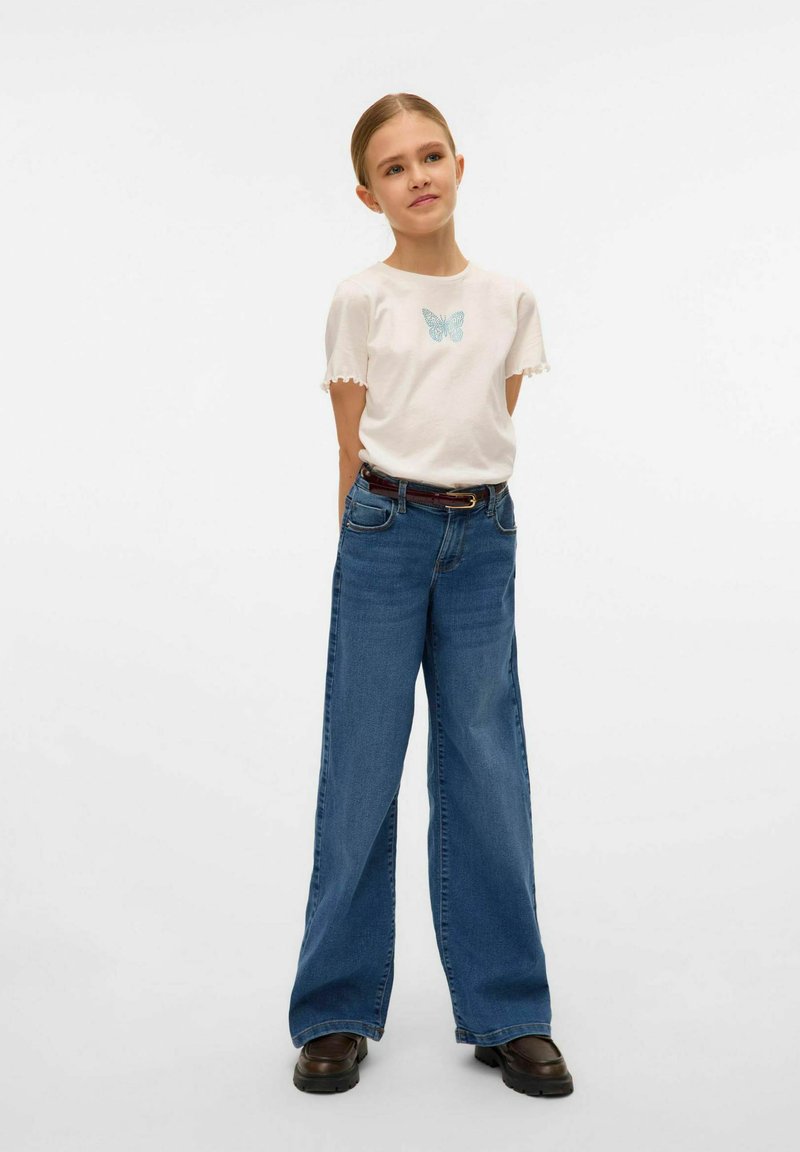 T-shirt blanc avec un motif de papillon bleu clair, manches à volants, associé à un jean bleu à jambes larges et des bottines noires.