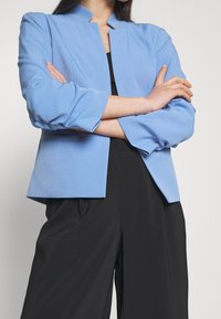 Svetle modrý blazer s moderním límcem, vytvarovaným střihem a rukávy do tří čtvrtin, zkombinovaný s černými širokými kalhotami.