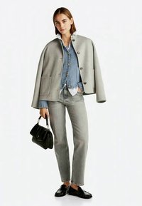 Grå ullcapejacka med fyra stora knappar, blå stickad tröja under, vit skjorta, grå jeans och svart handväska. Svarta loafers fulländar outfiten.