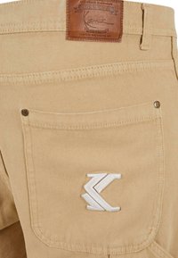 Beige kangasta valmistetut housut, joissa on tekstuuripinta. Takapocketissa on ruskea nahkalappu ja valkoinen brodeerattu logo. Mukana metalliset niitit.