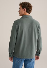 Világos zöld színű, galléros, hosszú ujjú ing, gombos mandzsettákkal és sima textúrával. Kék farmernadrággal párosítva.