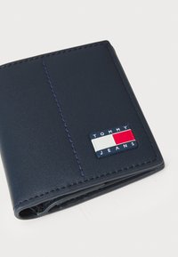 Carteira de couro azul-marinho com uma costura vertical, um patch de logótipo texturizado em azul, branco e vermelho, e um design fino, em bi-fold.