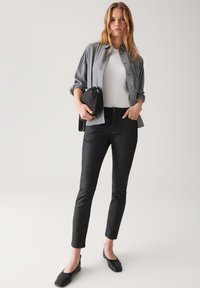 Giovane donna in piedi con una maglietta grigia sopra una maglietta bianca, pantaloni neri aderenti, ballerine nere e che tiene una piccola borsa nera all'altezza della vita.