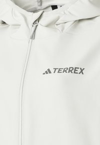 Veste softshell blanche avec capuche et fermeture éclair, avec un logo gris de la marque Terrex sur la poitrine.
