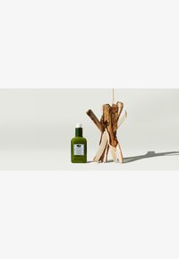 Bouteille en verre vert foncé avec un bouchon-pompe, étiquetée, à côté d'une structure en bois avec des bâtons de couleur claire. Design simple et épuré sur un fond pâle.