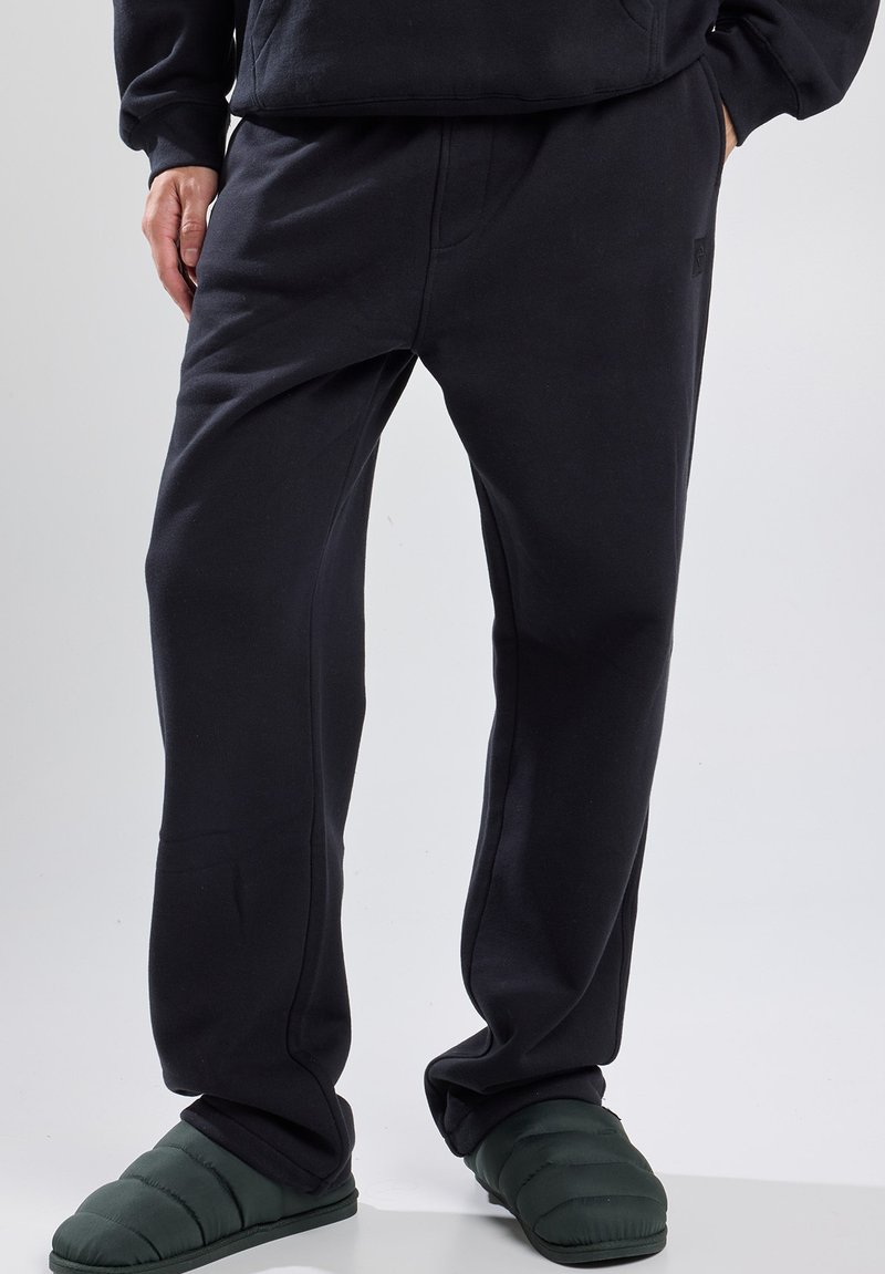 Pantalones de chándal negros hechos de tejido suave, con un corte relajado, cintura texturizada, bolsillos laterales y un discreto detalle de logo en la pierna.