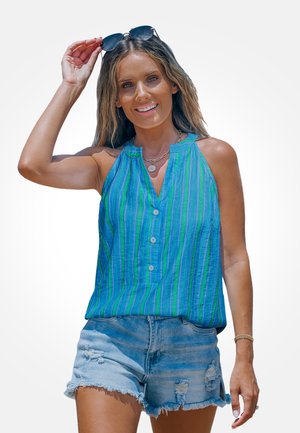 Frau lächelt und hebt ihre Sonnenbrille, trägt ein ärmelloses Top mit blauen und grünen Streifen sowie zerrissene Jeansshorts vor einem einfarbigen Hintergrund.