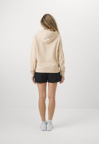 Felpa beige con una consistenza morbida e cappuccio, abbinata a pantaloni corti neri. L'outfit presenta una vestibilità rilassata, cuciture visibili e polsini elasticizzati.