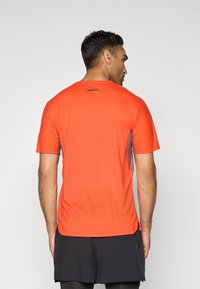Mand med ryggen til iført en lys orange perforeret kortærmet træningsskjorte og sorte shorts mod en ensfarvet baggrund.