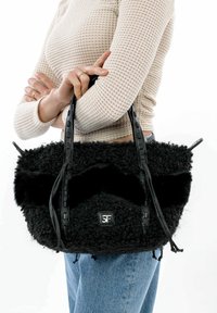 Melnas kažokādas tekstūras rokassoma ar gludiem ādas siksnām un logotipa uzšuvi. Iezīmē apaļu formu un izteiksmīgus pūkainus dizaina akcentus.