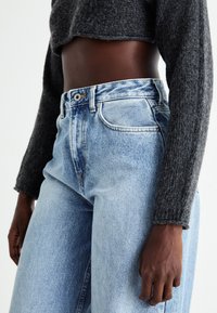 Blugi deschiși, cu talie înaltă, cu un design decolorat, cusături clare și patru buzunare, asortați cu un top cu mâneci lungi texturat de culoare gri închis.