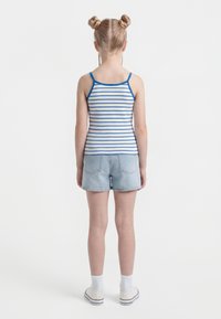 Girl with two braided buns wearing a blue and white striped tank top, light denim shorts, white socks, and white sneakers, standing with back to camera.