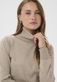 Pull à col roulé beige avec col et poignets côtelés. Texture douce, coupe ajustée. Bracelet chaîne en or et boucles d'oreilles créoles en accessoires.