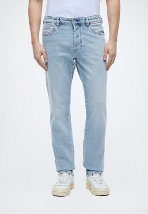Jean bleu clair à jambe droite porté avec des baskets blanches et un t-shirt blanc sur un fond blanc uni.