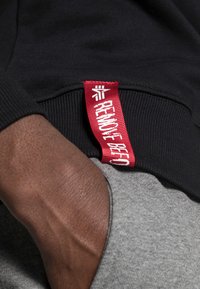 Zwarte sweatshirt met een rode tag met de tekst "VERWIJDER BAARD." Grijze accenten tonen textuurcontrast. Close-up van de stof en een hand.