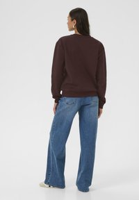 Sweatshirt marron à manches longues et poignets côtelés, associé à un jean bleu ample et des chaussures beiges. Fond uni.