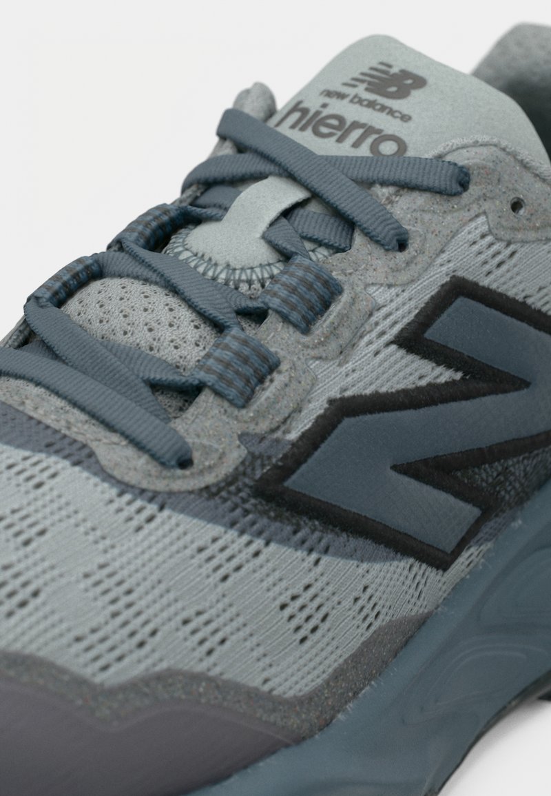 Chaussure de sport grise avec tige en mesh texturé, accents gris foncé, lacets bleus et logo noir proéminent. Dispose d'une semelle rembourrée et d'un embout renforcé.