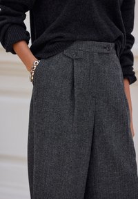 Graue Fischgrät-Hose mit tailliertem Schnitt, ausgestattet mit zwei vorderen Knöpfen und einer Seitentasche. Kombiniert mit einem dunklen Strickpullover.