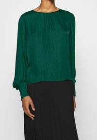 Blusa verde in tessuto leggero con maniche lunghe, scollatura tonda e dettagli plissettati nella parte anteriore. Indossata sopra una gonna nera.