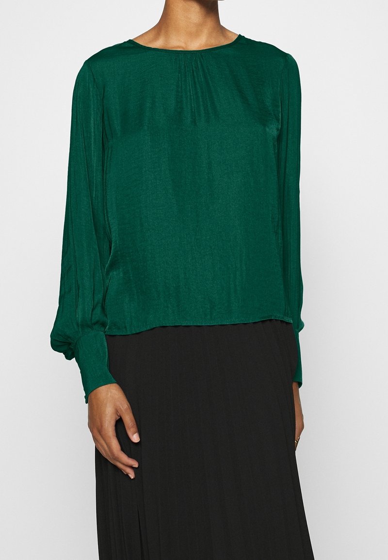 Blusa verde in tessuto leggero con maniche lunghe, scollatura tonda e dettagli plissettati nella parte anteriore. Indossata sopra una gonna nera.