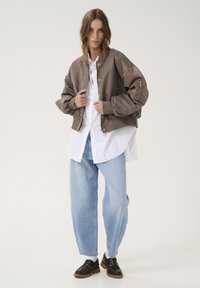 Brown satin bomber jacket with ribbed cuffs and zipper pocket, layered over a white button-up shirt and light blue baggy jeans.