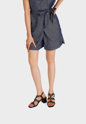 Donna che indossa pantaloncini blu scuro con cintura e sandali neri con tacco e punta aperta con dettagli circolari, in piedi su uno sfondo semplice.