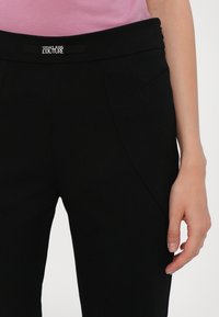 Črne hlače z gladko teksturo, ki imajo elastičen pas in nagnjene sprednje žepke. Na pasu je oznaka z logotipom Versace.